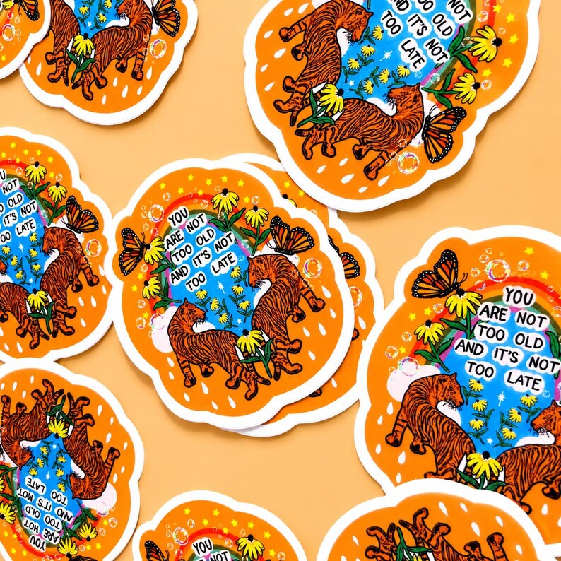 May include: Die-cut stickers featuring two orange tigers, yellow flowers, a butterfly, and a rainbow against a blue sky. The text reads, "YOU ARE NOT TOO OLD AND IT'S NOT TOO LATE." The stickers have an orange border with white raindrop shapes.