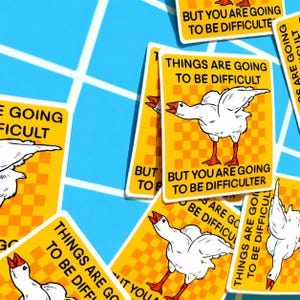 May include: A collection of yellow stickers with a white goose illustration and the text "THINGS ARE GOING TO BE DIFFICULT" and "BUT YOU ARE GOING TO BE DIFFICULTER". The stickers have a checkered background and are scattered on a blue surface.