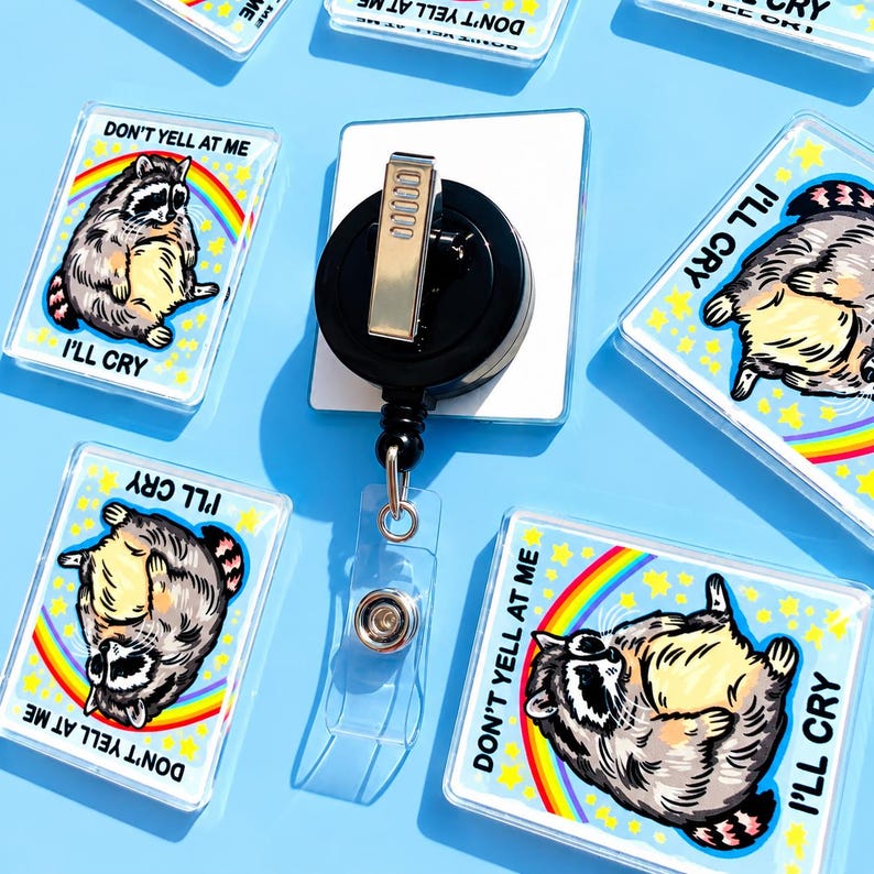 May include: A rectangular badge reel featuring a graphic of a raccoon with a rainbow and yellow stars, with the text DON'T YELL AT ME I'LL CRY. The badge reel has a black circular housing with a silver metal clip on the back and a clear strap.