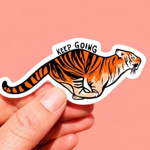 Puede incluir: Una pegatina troquelada que muestra un tigre naranja con rayas negras en posición de carrera. El tigre aparece de perfil mirando hacia la derecha. Sobre el tigre, el texto KEEP GOING está impreso en letras mayúsculas de color negro.