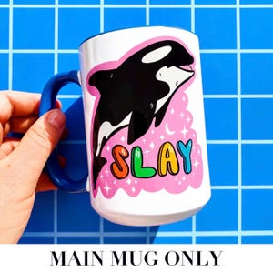 May include: White ceramic mug with a blue handle featuring a black and white orca illustration on a pink background. The word "SLAY" is written in colorful letters. The mug is held against a blue grid background.