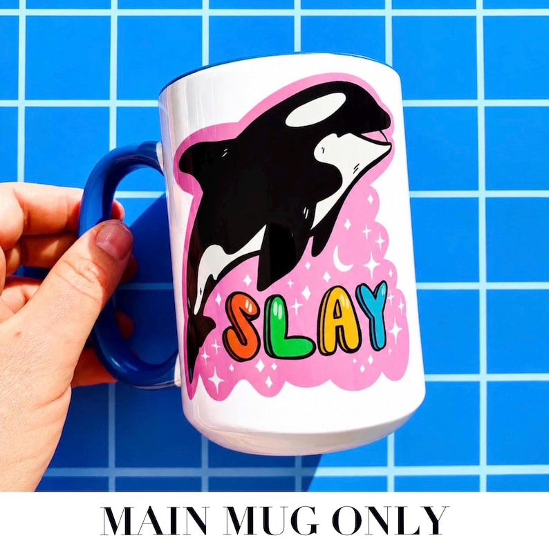 May include: White ceramic mug with a blue handle featuring a black and white orca illustration on a pink background. The word "SLAY" is written in colorful letters. The mug is held against a blue grid background.