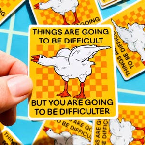 May include: Square yellow sticker with a white goose illustration and the text "THINGS ARE GOING TO BE DIFFICULT BUT YOU ARE GOING TO BE DIFFICULTER". The goose has orange feet and beak, and is set against a checkered background.