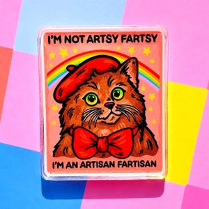 May include: A rectangular sticker featuring a cartoon cat wearing a red beret and bow tie. The cat is brown with green eyes and a white mustache. The sticker has the text "I'M NOT ARTSY FARTSY" and "I'M AN ARTISAN FARTISAN" with a rainbow and stars.