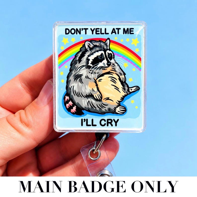 May include: A square badge reel featuring a cartoon raccoon with grey fur and a cream-colored belly. The background is light blue with a colorful rainbow and yellow stars. The text reads DON'T YELL AT ME I'LL CRY in bold black capital letters.