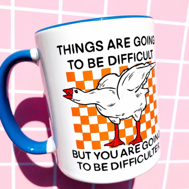May include: White ceramic mug with a blue handle and rim. The mug features a cartoon goose illustration on an orange and white checkered background. Text on the mug reads "THINGS ARE GOING TO BE DIFFICULT" and "BUT YOU ARE GOING TO BE DIFFICULT-ER."
