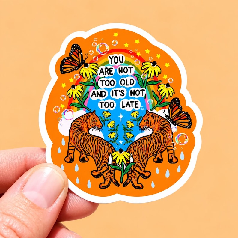 May include: A die-cut sticker with an orange background featuring two orange and black tigers facing each other, yellow flowers, monarch butterflies, a rainbow, bubbles, and raindrops. The text reads: YOU ARE NOT TOO OLD AND IT'S NOT TOO LATE.