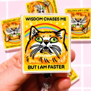 May include: A rectangular badge reel featuring a yellow background with a graphic illustration of an orange cat with wide green eyes. Above the cat, the text reads WISDOM CHASES ME, and below, BUT I AM FASTER. A colorful rainbow arches behind the cat.