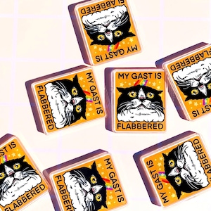 Puede incluir: Un conjunto de imanes rectangulares con una ilustraci&oacute;n de gato de dibujos animados. El gato es blanco y negro con ojos amarillos y un arco&iacute;ris. Los imanes tienen el texto "MY GAST IS FLABBERED" sobre un fondo amarillo con estrellas blancas.