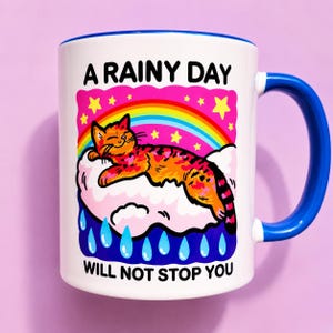May include: A white ceramic mug with a blue handle and rim, featuring a graphic of an orange tabby cat sleeping on a pink cloud under a rainbow and yellow stars, with blue raindrops below and the text A RAINY DAY WILL NOT STOP YOU.
