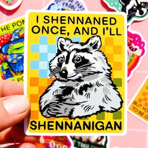 Puede incluir: Una pegatina cuadrada con una ilustración de mapache y el texto "I SHENNANED ONCE, AND I'LL SHENNANIGAN" sobre un fondo amarillo con un patrón de cuadros. El mapache es negro, blanco y gris.