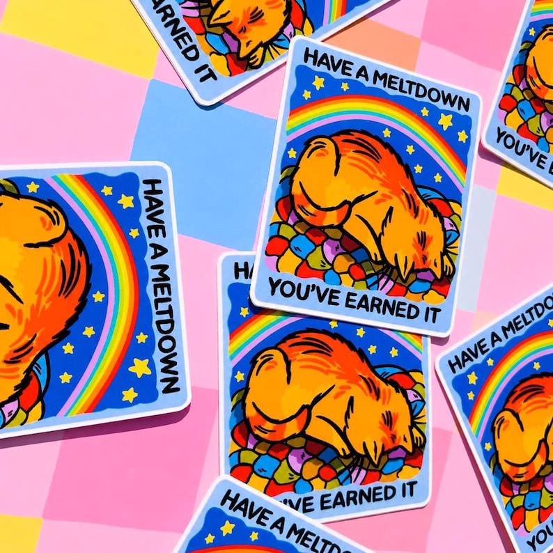 Puede incluir: Conjunto de pegatinas con una ilustraci&oacute;n de gato de dibujos animados. Cada pegatina presenta un fondo azul con un arco&iacute;ris, estrellas y las frases "Have a Meltdown" y "You've Earned It". El gato es naranja y est&aacute; tumbado sobre formas coloridas.