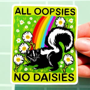 May include: A rectangular sticker featuring a black and white skunk standing in a bright green field with white daisies. A colourful rainbow arches behind the skunk. The text ALL OOPSIES NO DAISIES is printed in black at the top and bottom edges.