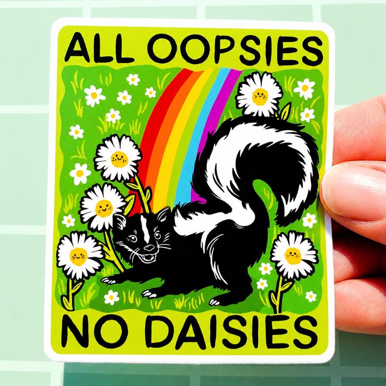 May include: A rectangular sticker featuring a black and white skunk standing in a bright green field with white daisies. A colourful rainbow arches behind the skunk. The text ALL OOPSIES NO DAISIES is printed in black at the top and bottom edges.