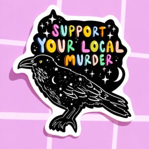May include: A sticker featuring a black raven with a starry design and the text "SUPPORT YOUR LOCAL MURDER" in colorful bubble letters. The sticker has a white border and is set against a pink background.