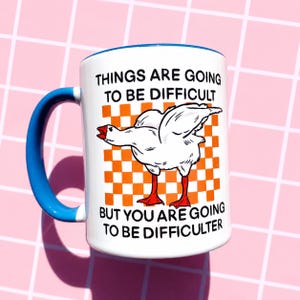 May include: White ceramic mug with a blue handle and rim. The mug features a cartoon goose illustration with the text "THINGS ARE GOING TO BE DIFFICULT BUT YOU ARE GOING TO BE DIFFICULTER" on a checkered orange background.