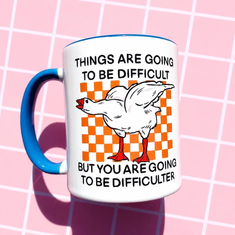 May include: White ceramic mug with a blue handle and rim. The mug features a cartoon goose illustration with the text "THINGS ARE GOING TO BE DIFFICULT BUT YOU ARE GOING TO BE DIFFICULTER" on a checkered orange background.
