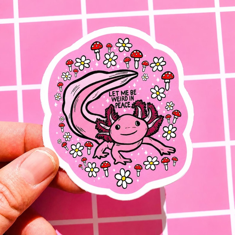 May include: A pink sticker featuring a cartoon axolotl with pink gills and a long tail, surrounded by white flowers and red-capped mushrooms. The sticker has the text "LET ME BE WEIRD IN PEACE" in black. The sticker is held by a hand.