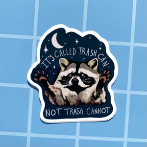 May include: A blue and white sticker with a raccoon in the center. The raccoon is holding up its paw and has a surprised expression. The text on the sticker reads "It's called trash can not trash cannot".