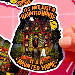 May include: A Halloween-themed sticker featuring a spooky haunted house illustration. The house is green and black, with glowing orange windows and is surrounded by pumpkins, ghosts, and a full moon. Text reads "It's Not Just a Haunted House, It's a Haunted Home."