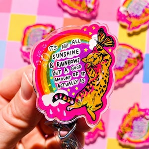 May include: A pink acrylic badge reel featuring a cartoon orange tabby cat reaching for a butterfly. A rainbow and clouds are in the background with the text: IT'S NOT ALL SUNSHINE & RAINBOWS BUT A GOOD AMOUNT OF IT ACTUALLY IS. The design includes yellow stars.