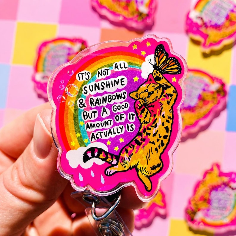 May include: A pink acrylic badge reel featuring a cartoon orange tabby cat reaching for a butterfly. A rainbow and clouds are in the background with the text: IT'S NOT ALL SUNSHINE & RAINBOWS BUT A GOOD AMOUNT OF IT ACTUALLY IS. The design includes yellow stars.
