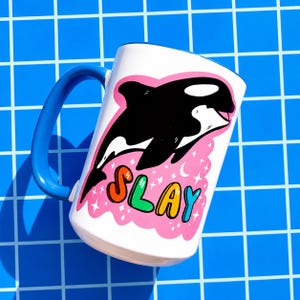 May include: White ceramic mug with a blue handle featuring a black and white orca illustration. The mug has the word "SLAY" in colorful letters on a pink cloud background. The mug is set against a blue grid background.