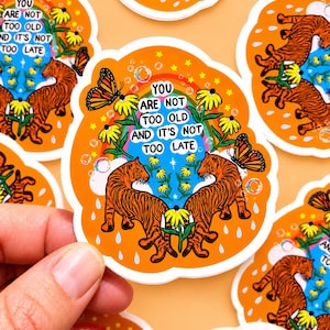 May include: A die-cut sticker with an orange background featuring two orange and black tigers facing each other. The design includes yellow flowers, monarch butterflies, a rainbow, bubbles, and the text: YOU ARE NOT TOO OLD AND IT'S NOT TOO LATE.