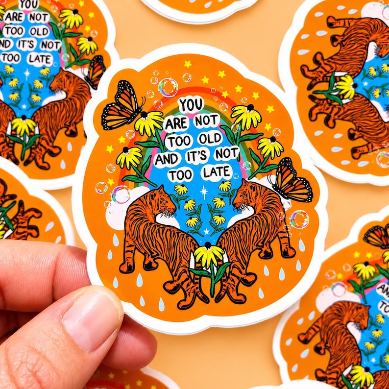May include: A die-cut sticker with an orange background featuring two orange and black tigers facing each other. The design includes yellow flowers, monarch butterflies, a rainbow, bubbles, and the text: YOU ARE NOT TOO OLD AND IT'S NOT TOO LATE.