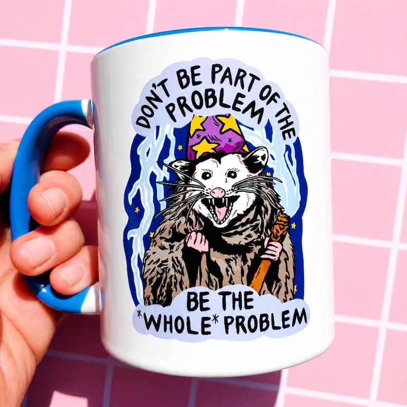 May include: White ceramic mug with a blue handle and rim. The mug features a cartoon opossum wearing a wizard hat and holding a staff, with the text "DON'T BE PART OF THE PROBLEM BE THE *WHOLE* PROBLEM."