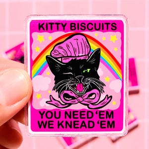 May include: A rectangular acrylic pin featuring a black cat wearing a pink chef hat and a pink bow. The background is pink with a rainbow, yellow stars, and the text KITTY BISCUITS YOU NEED 'EM WE KNEAD 'EM in bold black capital letters.