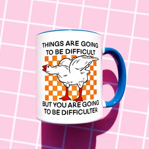 May include: White ceramic mug with a blue handle and rim. The mug features a cartoon goose illustration on an orange and white checkered background. Text on the mug reads "THINGS ARE GOING TO BE DIFFICULT BUT YOU ARE GOING TO BE DIFFICULTER."