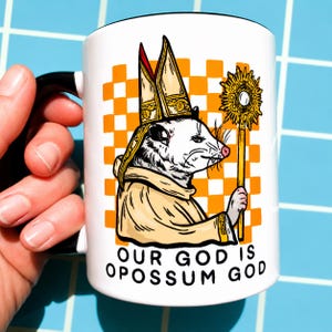 Könnte beinhalten: Weiße Keramik-Tasse mit schwarzem Henkel. Sie zeigt ein Cartoon-Opossum in religiöser Kleidung, das einen goldenen Stab hält, mit dem Text "OUR GOD IS OPOSSUM GOD". Der Hintergrund hat ein orange-weißes Schachbrettmuster.