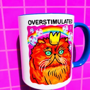 K&ouml;nnte beinhalten: Ein wei&szlig;er Keramikbecher mit blauem Henkel und einer Grafik einer orangefarbenen Katze mit gelber Krone. &Uuml;ber der Katze befinden sich ein Regenbogen und Seifenblasen, sowie der Text OVERSTIMULATED in schwarzen Gro&szlig;buchstaben oben.