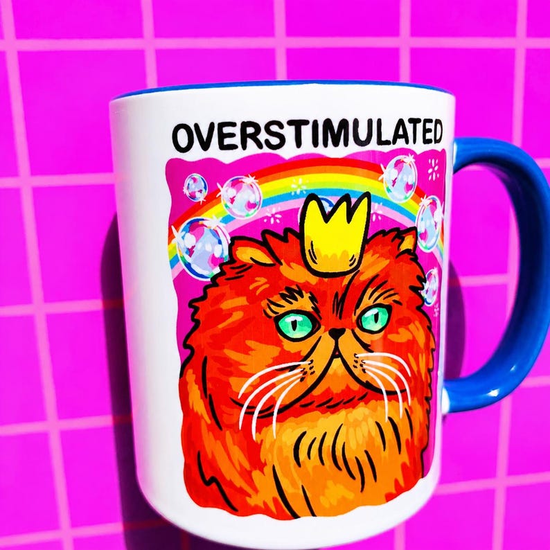 K&ouml;nnte beinhalten: Ein wei&szlig;er Keramikbecher mit blauem Henkel und einer Grafik einer orangefarbenen Katze mit gelber Krone. &Uuml;ber der Katze befinden sich ein Regenbogen und Seifenblasen, sowie der Text OVERSTIMULATED in schwarzen Gro&szlig;buchstaben oben.