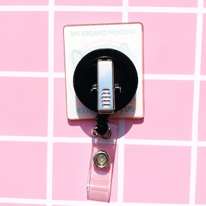 May include: A square badge reel with a black circular housing and a silver metal clip on the back. The badge reel features a clear plastic strap with a silver snap button. The text WISDOM CHASES ME is printed on the square base of the badge reel.