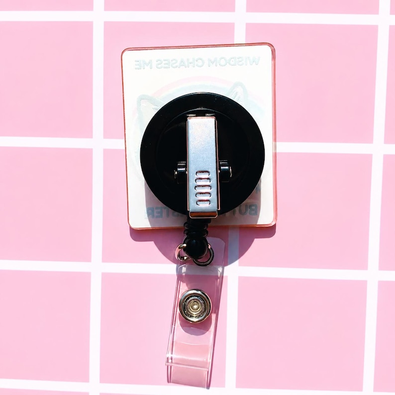 May include: A square badge reel with a black circular housing and a silver metal clip on the back. The badge reel features a clear plastic strap with a silver snap button. The text WISDOM CHASES ME is printed on the square base of the badge reel.