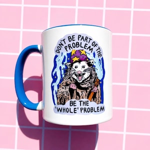 Pu&ograve; includere: Tazza in ceramica bianca con manico e bordo blu. La tazza presenta un opossum dei cartoni animati con un cappello da mago e un bastone, con il testo "DON'T BE PART OF THE PROBLEM BE THE *WHOLE* PROBLEM."