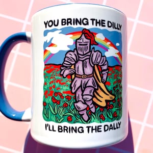 May include: White ceramic mug with a blue handle and rim. The mug features a cartoon knight in armor running through a field of red flowers under a rainbow. Text on the mug reads "YOU BRING THE DILLY, I'LL BRING THE DALLY."