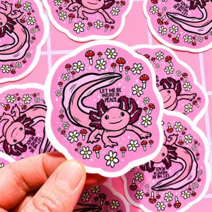 May include: Pink axolotl sticker with the text "LET ME BE WEIRD IN PEACE". The sticker features a cartoon axolotl surrounded by white daisies and red mushroom illustrations. The sticker is held by a hand.
