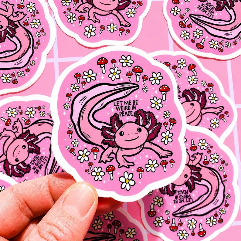May include: Pink axolotl sticker with the text "LET ME BE WEIRD IN PEACE". The sticker features a cartoon axolotl surrounded by white daisies and red mushroom illustrations. The sticker is held by a hand.