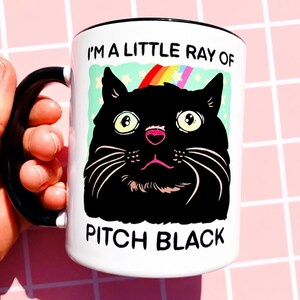 May include: White ceramic mug with a black handle and rim. The mug features a cartoon illustration of a black cat with a rainbow and the text "I'M A LITTLE RAY OF PITCH BLACK". The cat has large yellow eyes and a pink nose.
