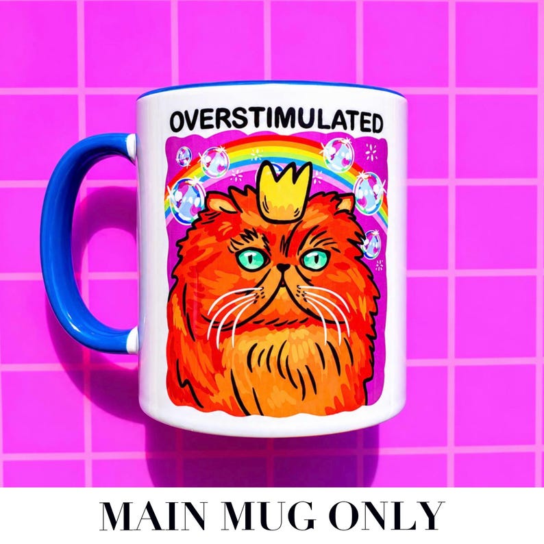 K&ouml;nnte beinhalten: Ein wei&szlig;er Keramikbecher mit blauem Henkel und einer Grafik einer flauschigen orangefarbenen Katze mit gelber Krone. &Uuml;ber der Katze steht der Text OVERSTIMULATED in schwarzen Buchstaben. Das Motiv zeigt einen bunten Regenbogen und Seifenblasen.