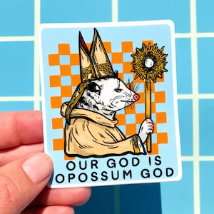May include: A sticker featuring an opossum dressed in religious garb, holding a golden staff with a sunburst design. The text "OUR GOD IS OPOSSUM GOD" is printed below the opossum on a light blue background with an orange checkered pattern.