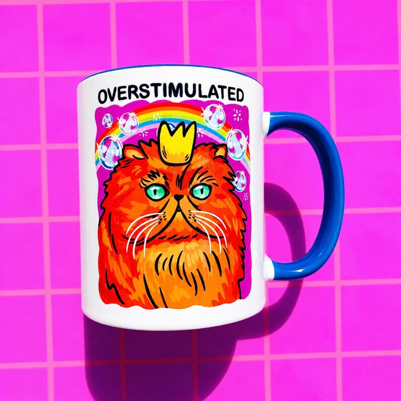 K&ouml;nnte beinhalten: Ein wei&szlig;er Keramikbecher mit blauem Henkel und einer Grafik einer orangefarbenen Katze mit gelber Krone. &Uuml;ber der Katze befinden sich ein Regenbogen und Seifenblasen, dar&uuml;ber steht der Text OVERSTIMULATED in schwarzen Gro&szlig;buchstaben.