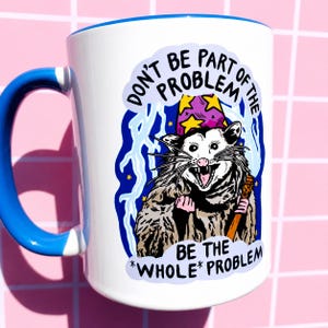 Pu&ograve; includere: Tazza in ceramica bianca con manico e bordo blu. La tazza presenta un opossum dei cartoni animati con un cappello da mago e un bastone. Il testo sulla tazza recita "DON'T BE PART OF THE PROBLEM BE THE *WHOLE* PROBLEM."
