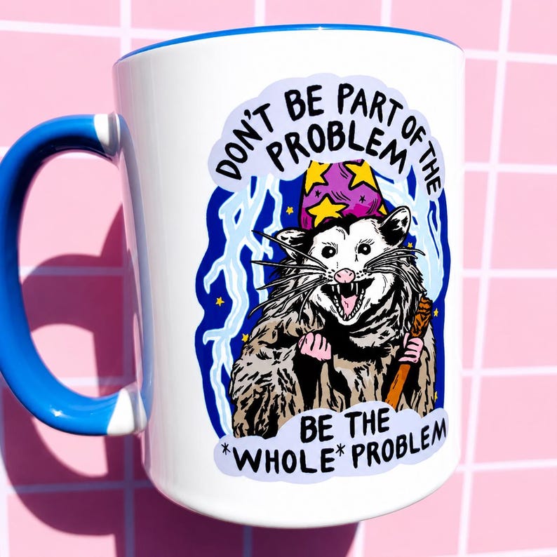 Pu&ograve; includere: Tazza in ceramica bianca con manico e bordo blu. La tazza presenta un opossum dei cartoni animati con un cappello da mago e un bastone. Il testo sulla tazza recita "DON'T BE PART OF THE PROBLEM BE THE *WHOLE* PROBLEM."