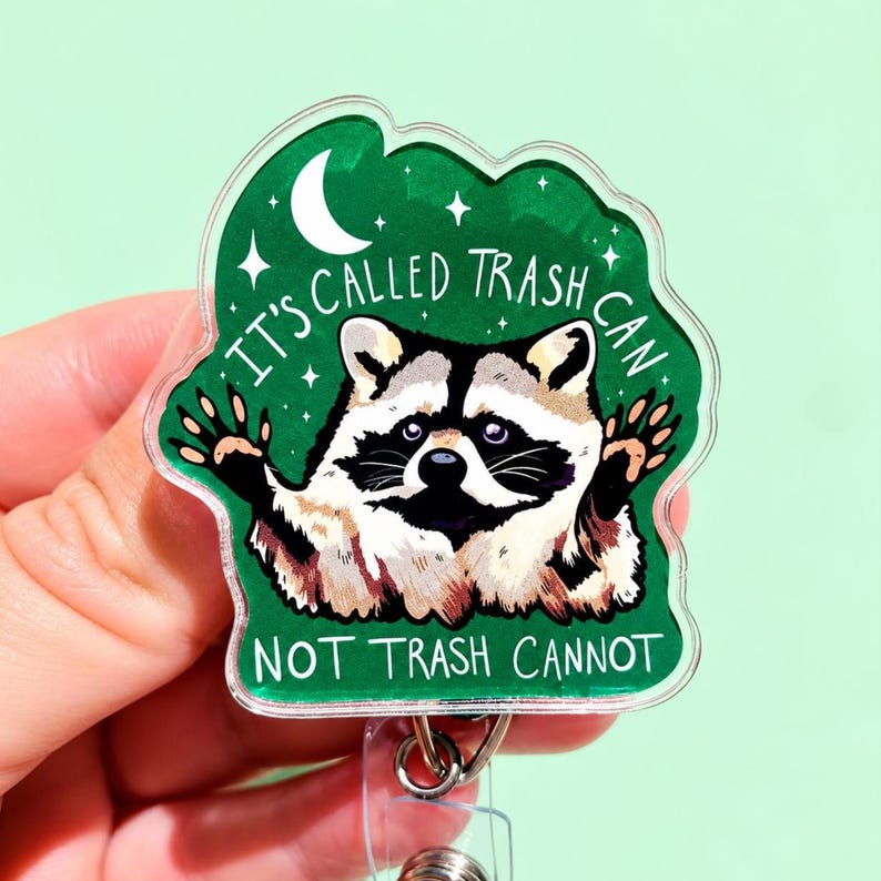 May include: An acrylic badge reel featuring a raccoon illustration on a green background with a white crescent moon and stars. The text reads, "IT'S CALLED TRASH CAN NOT TRASH CANNOT" in white lettering. The design is encased in clear acrylic.