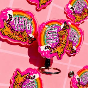 May include: A collection of acrylic badge reels featuring an orange tabby cat reaching for a butterfly. The design includes a rainbow, clouds, and the text: IT'S NOT ALL SUNSHINE & RAINBOWS BUT A GOOD AMOUNT OF IT ACTUALLY IS, set against a pink background.