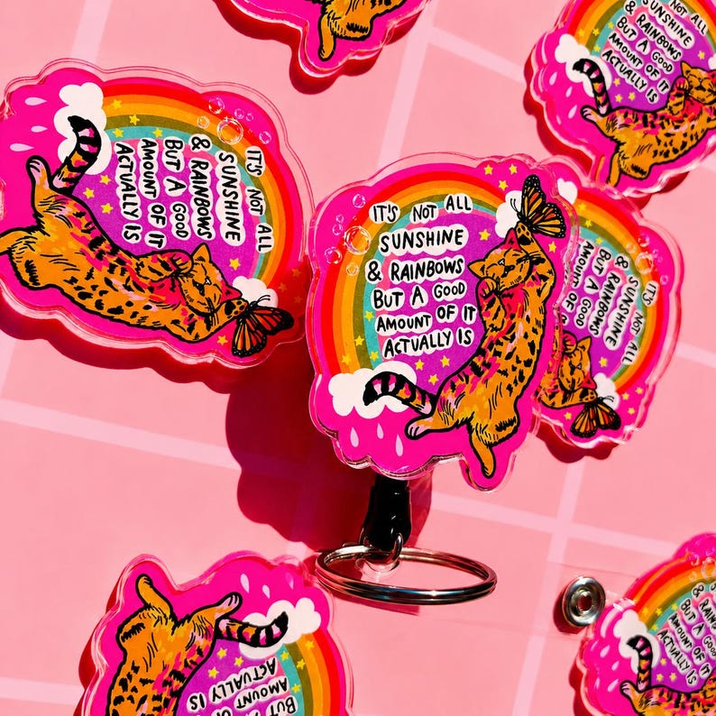 May include: A collection of acrylic badge reels featuring an orange tabby cat reaching for a butterfly. The design includes a rainbow, clouds, and the text: IT'S NOT ALL SUNSHINE & RAINBOWS BUT A GOOD AMOUNT OF IT ACTUALLY IS, set against a pink background.